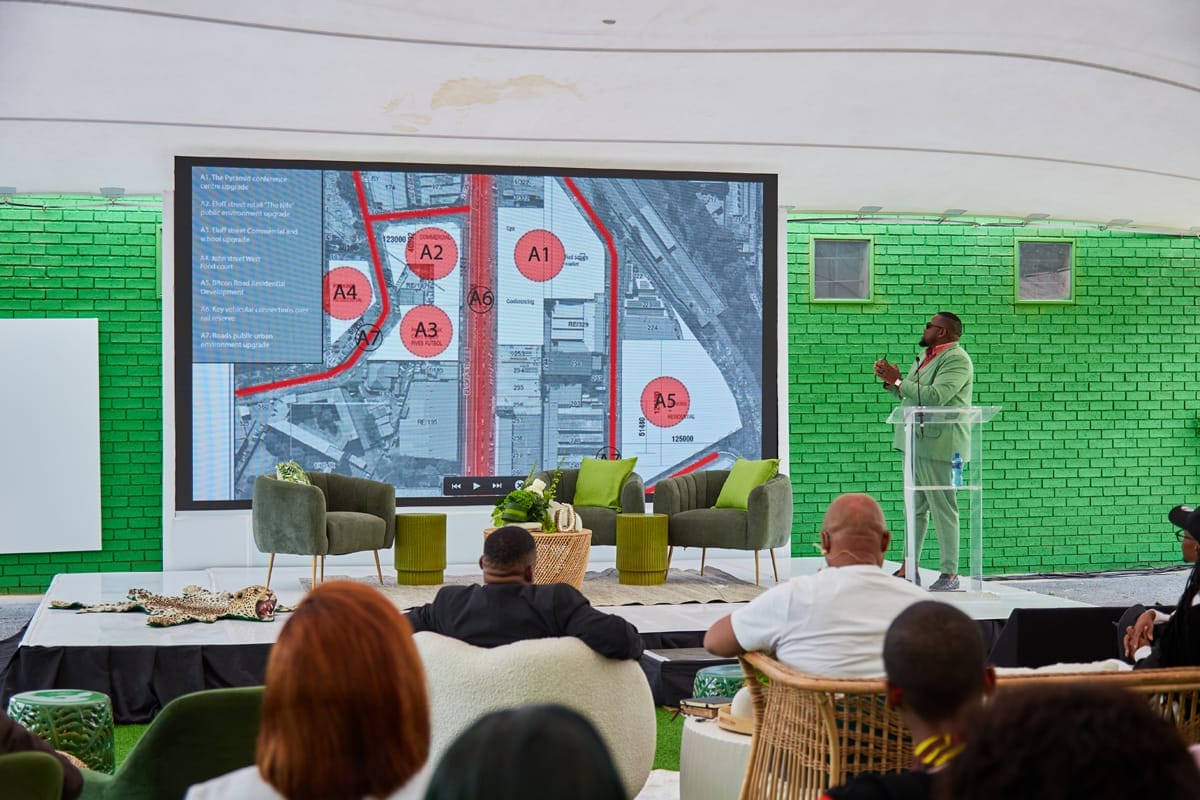 A presenter in a light green suit speaks at a clear lectern beside a large screen showing a site map with labelled red circles. The audience sits on modern chairs, and the background features a bright green brick wall.
