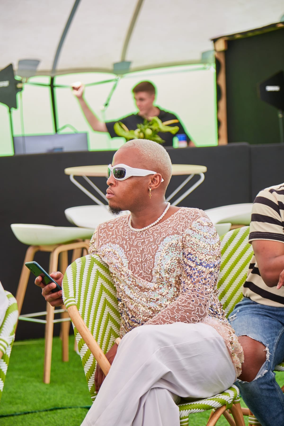 A person with short blonde hair, wearing white sunglasses, a sheer sequinned top, and white trousers, sits on a green patterned chair holding a mobile. A DJ is in the background at a booth with green lighting. Another person sits partially visible beside them.