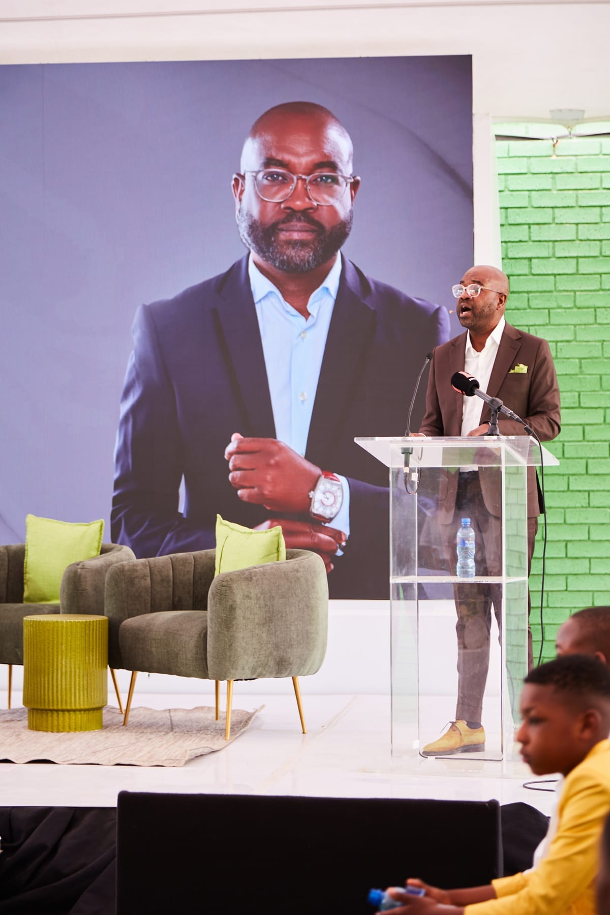 A man in a dark suit stands at a clear lectern, speaking, with a water bottle on the lectern. Behind him is a large portrait of himself. Green chairs, a small table, and green brick walls are visible on stage. Members of the audience are seen in the foreground.