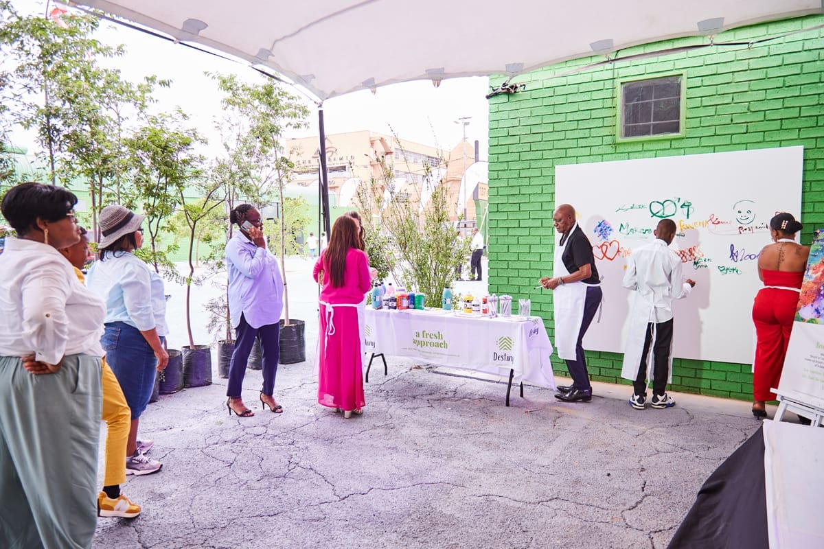 Several people stand and watch as others paint colourful designs and words on a large white board attached to a bright green wall outdoors. A table with art supplies and a “fresh approach” sign is in front of the artists. Trees and buildings are visible in the background.