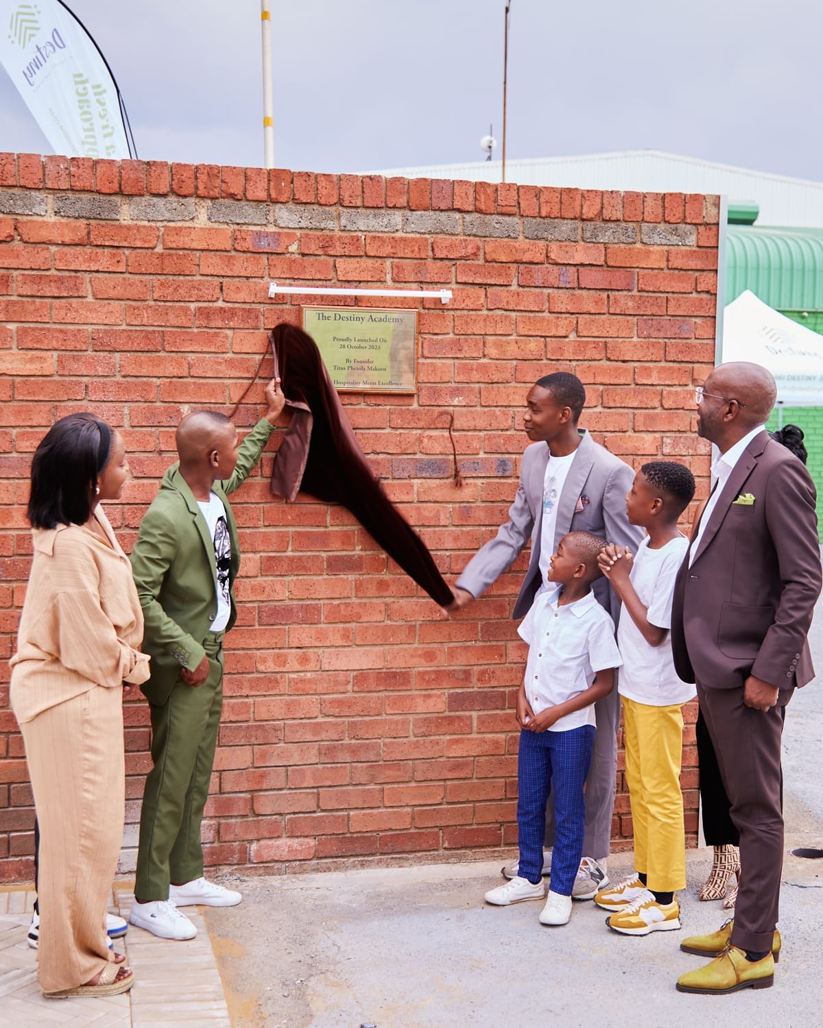 Five people, including three adults and two children, stand by a red brick wall as a brown cloth is being lifted to unveil a gold plaque. They are dressed in smart-casual clothes, and all are looking at the plaque. A green banner is visible in the background.