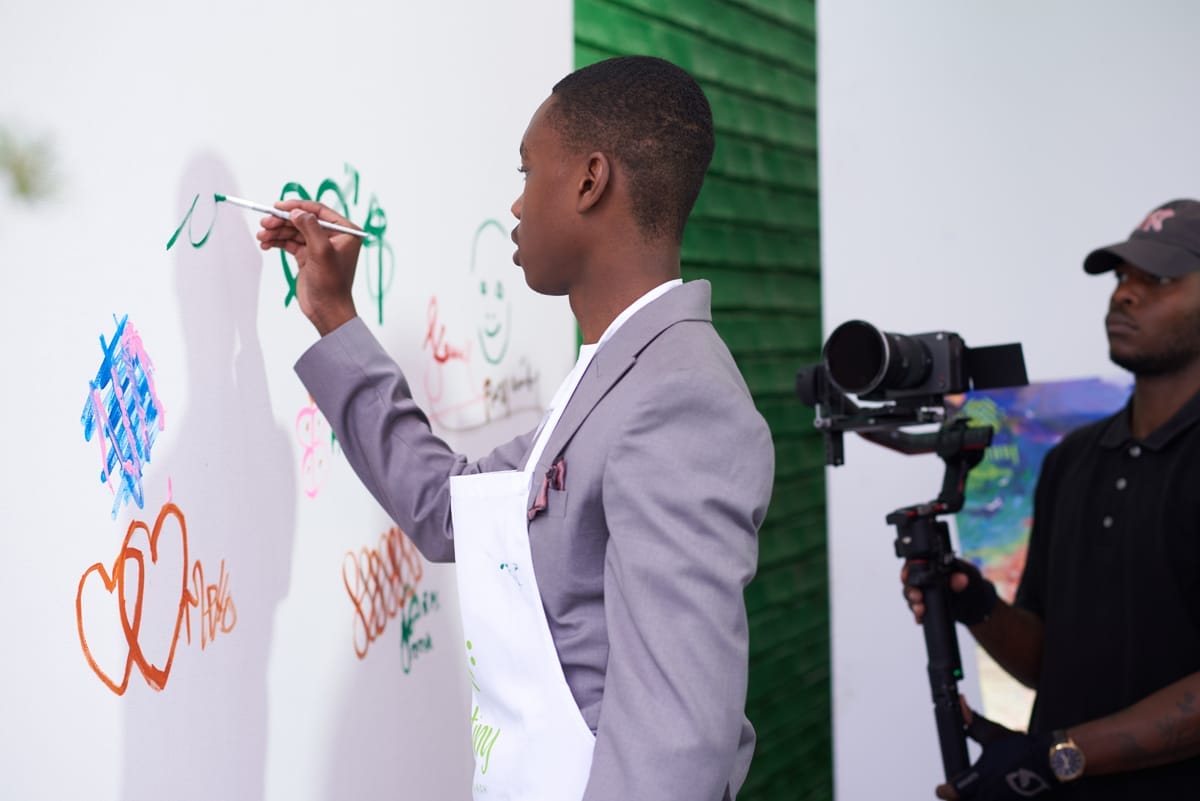 A young man in a light grey suit and white apron paints colourful graffiti and doodles on a white wall. Another person wearing black holds a camera on a stabiliser, filming him. The background features a green, textured wall panel.