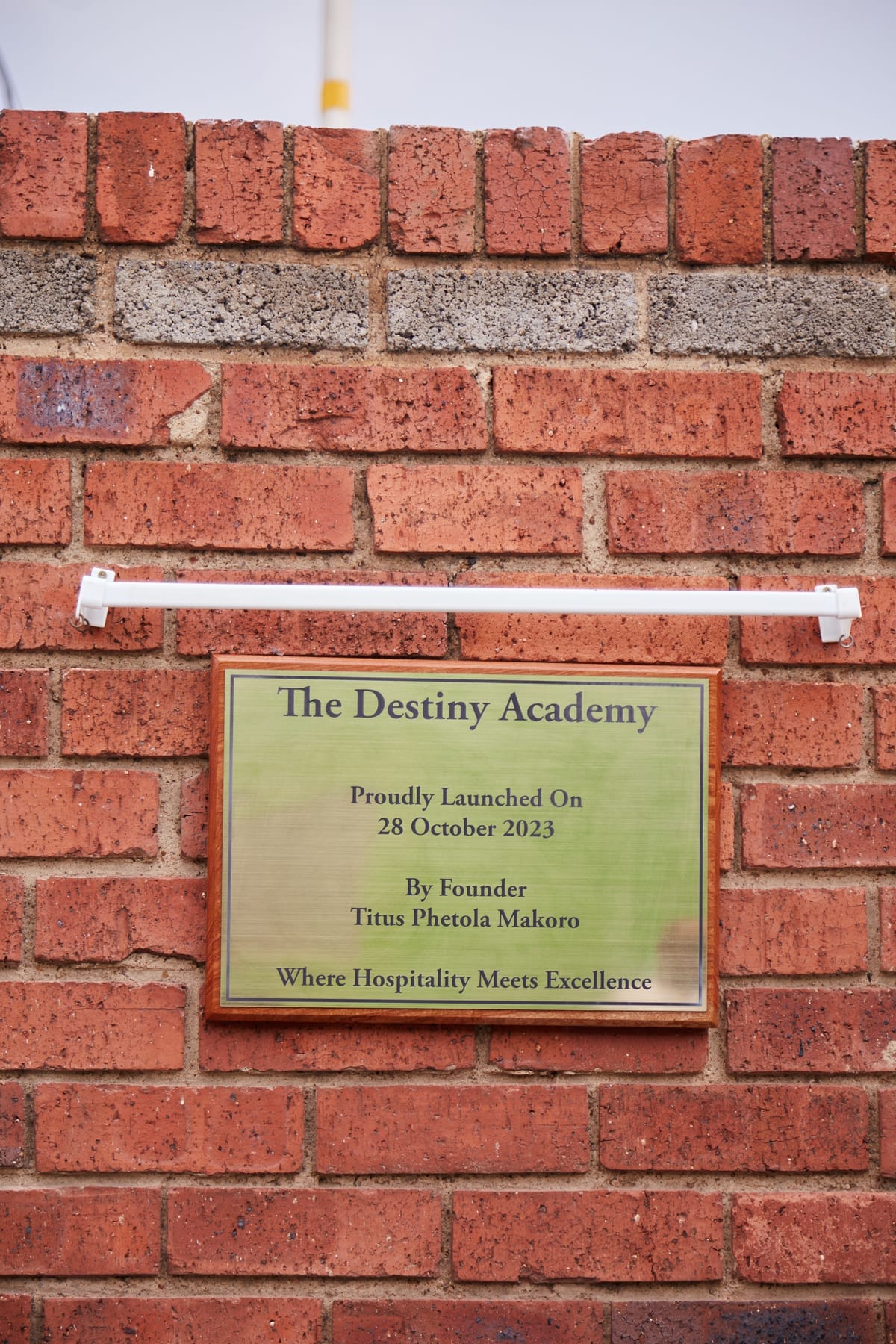 A brass plaque mounted on a red brick wall reads: “The Destiny Academy. Proudly Launched On 28 October 2023. By Founder Titus Phetola Makoro. Where Hospitality Meets Excellence.” A white fitting is installed above the plaque.