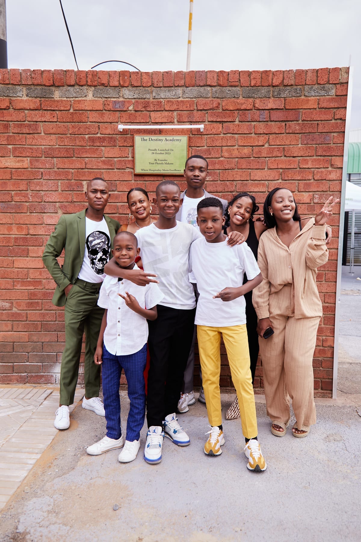 Eight smiling people, including children and adults, stand close together in front of a red brick wall with a yellow plaque. They wear casual and semi-formal clothes in bright colours. The group appears happy and relaxed. The scene is outdoors, on a paved surface.