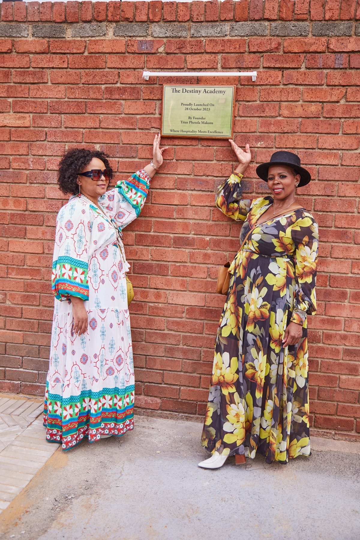 Two women stand smiling against a brick wall. Both wear long, colourful dresses and sunglasses; one wears a black hat. They each point up to a plaque on the wall that reads 