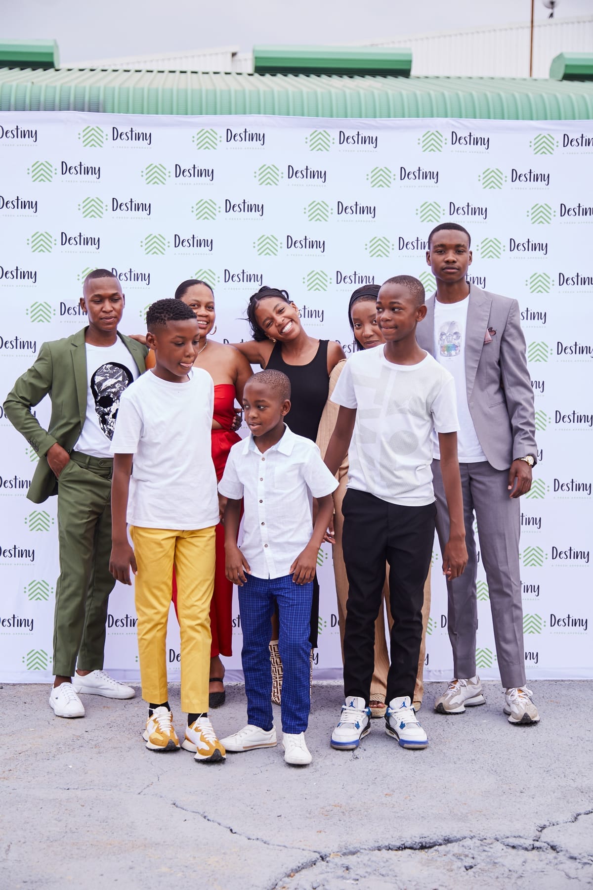 A group of seven smiling youths and teens, dressed in colourful semi-formal outfits, pose together in front of a white 