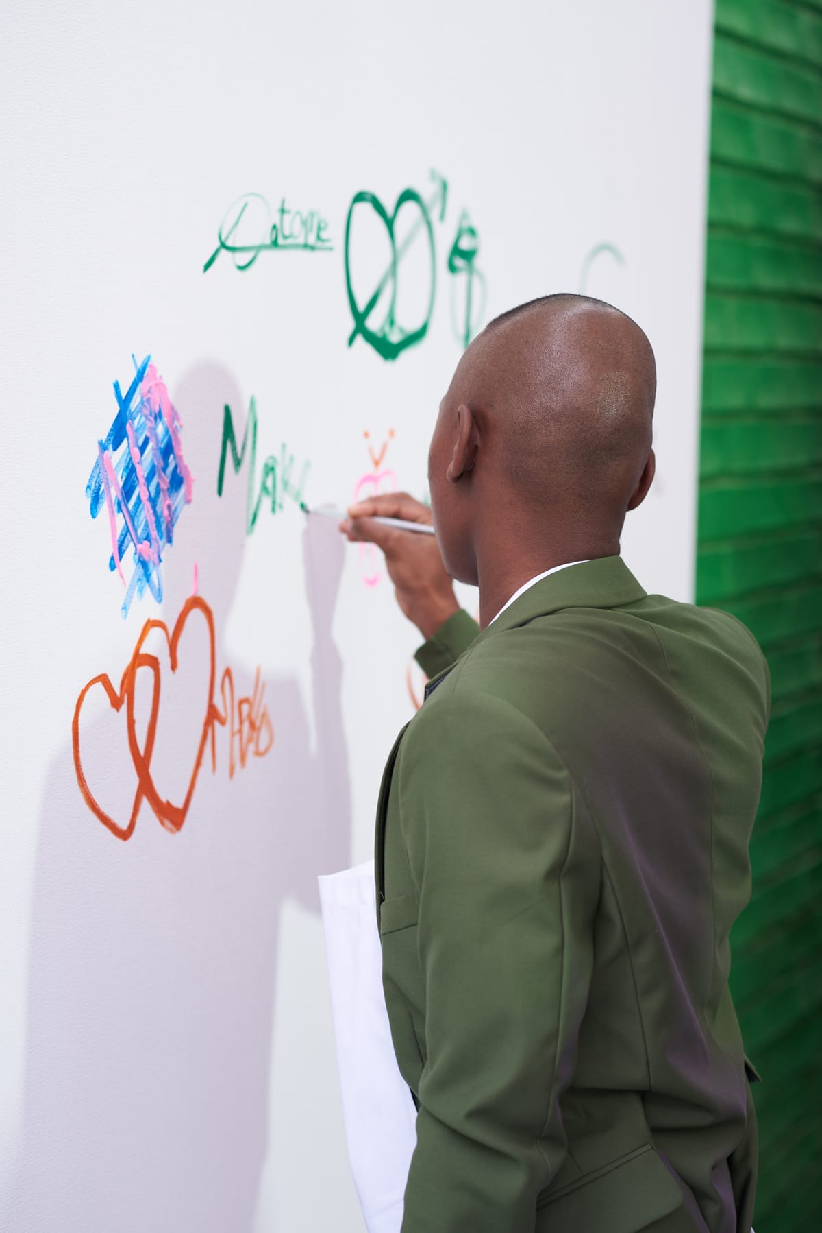 A person in a green suit writes colourful words and doodles, including hearts and hashtags, on a white wall. The person’s back faces the camera, and they hold a white tote bag. A green, textured wall is visible on the right side.