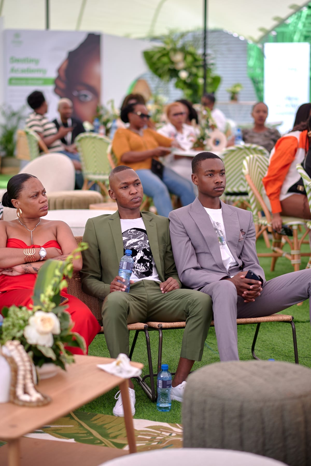 Three people sit on a sofa at an indoor event. The woman on the left wears a red dress, the man in the middle wears a green suit, and the man on the right wears a grey suit. Other attendees and green décor are visible in the background.