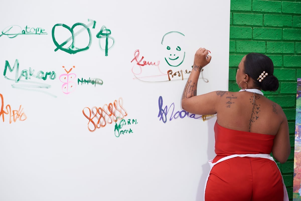 A woman in a red outfit with tattoos on her back writes on a white wall using markers. The wall displays colourful drawings, signatures, and doodles. She stands in front of a green brick wall, her hair tied back and secured with pearl hairpins.