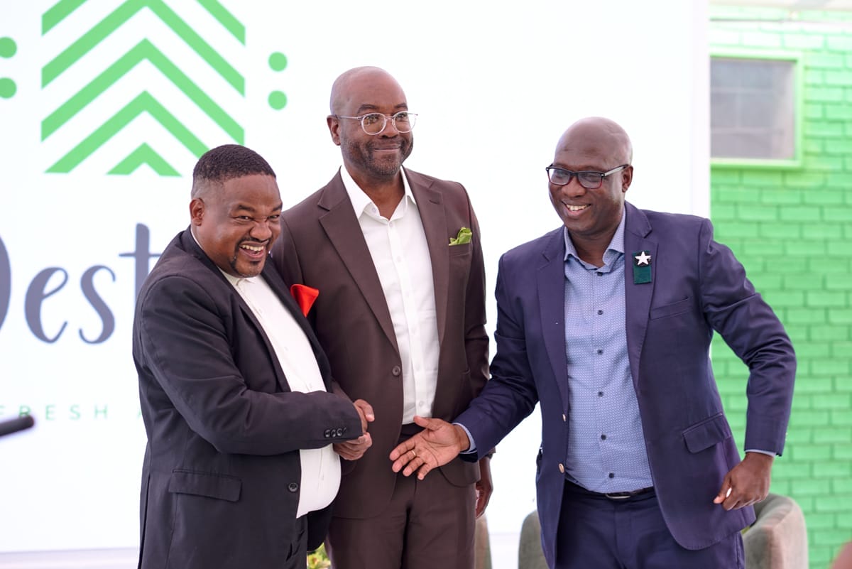 Three men in suits smile and shake hands in front of a white backdrop with green geometric designs and text. One man wears a black suit with a red pocket square, another wears a brown suit, and the third wears a blue suit with glasses and a star pin.