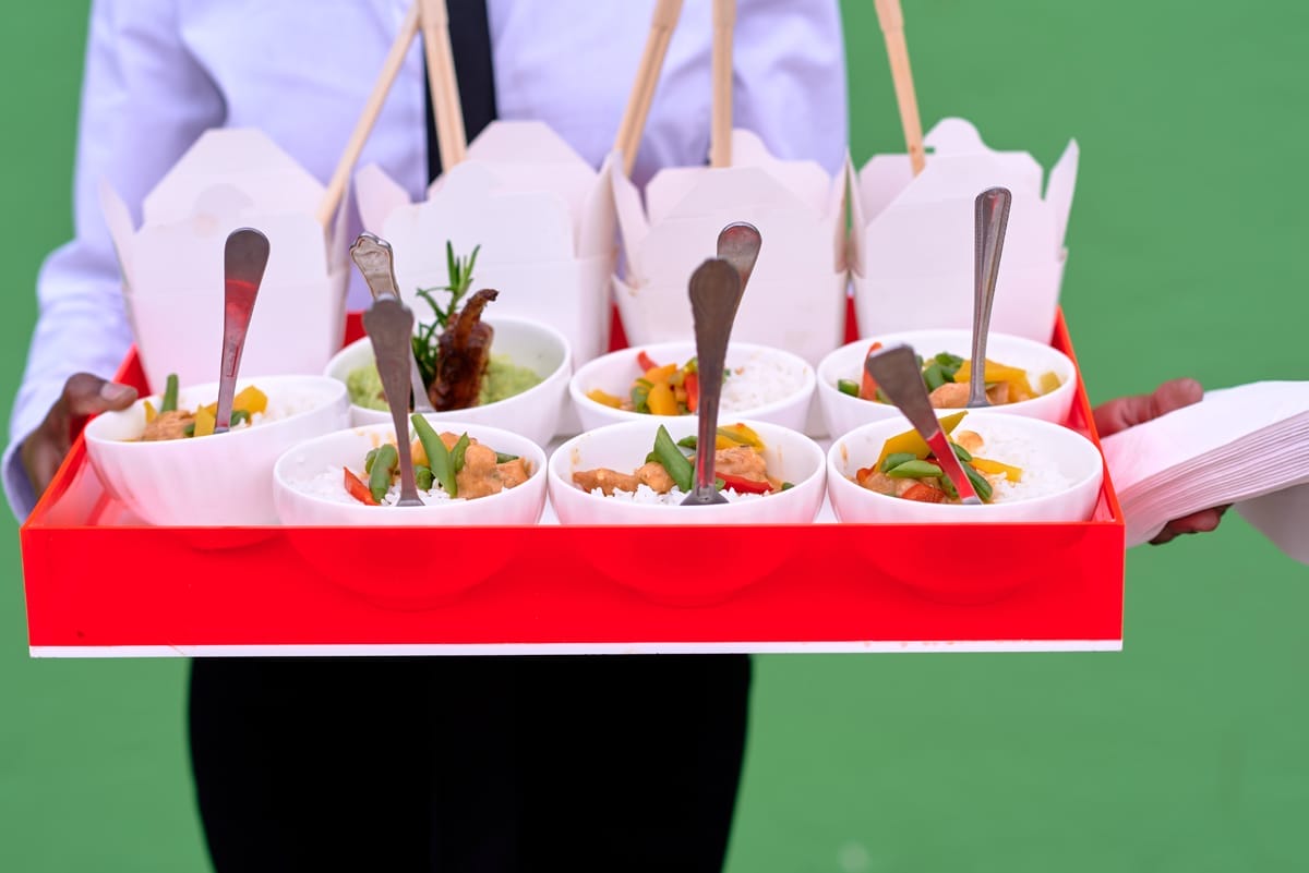 A person in a white shirt carries a red tray with eight white bowls of rice topped with colourful vegetables and herbs, each with a fork. Behind the bowls are white takeaway boxes with skewers. The background is green and out of focus.