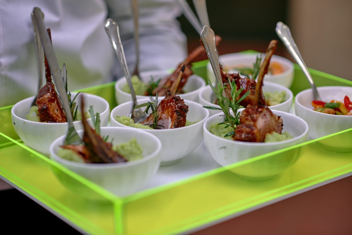 A waiter carries a neon green tray holding small white bowls. Each bowl contains two glazed lamb cutlets, a sprig of rosemary, and a green purée, with a spoon resting inside. Another bowl at the back holds a colourful vegetable salad.