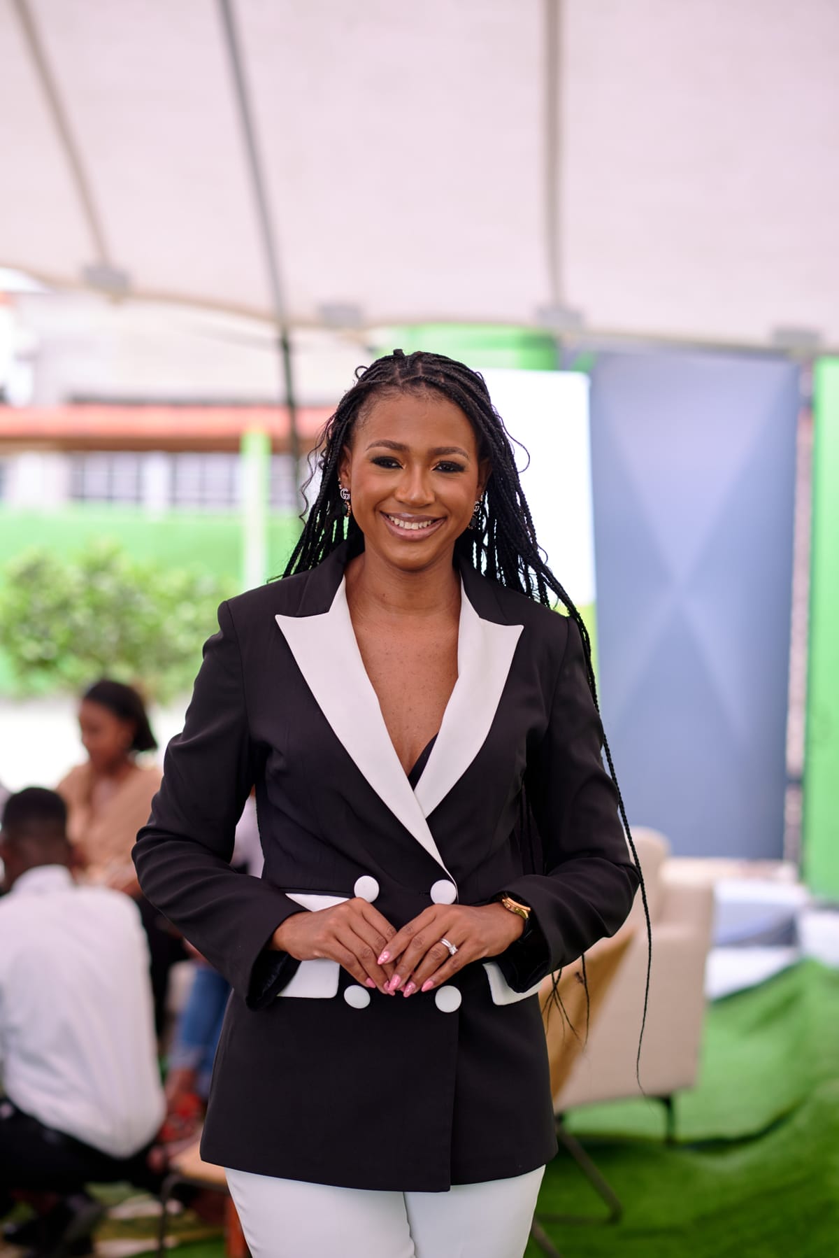 A woman with long plaited hair stands smiling under a marquee. She wears a black blazer with white lapels and buttons, and white trousers. Her hands are clasped in front. In the background, people sit and talk near green and blue decor elements.