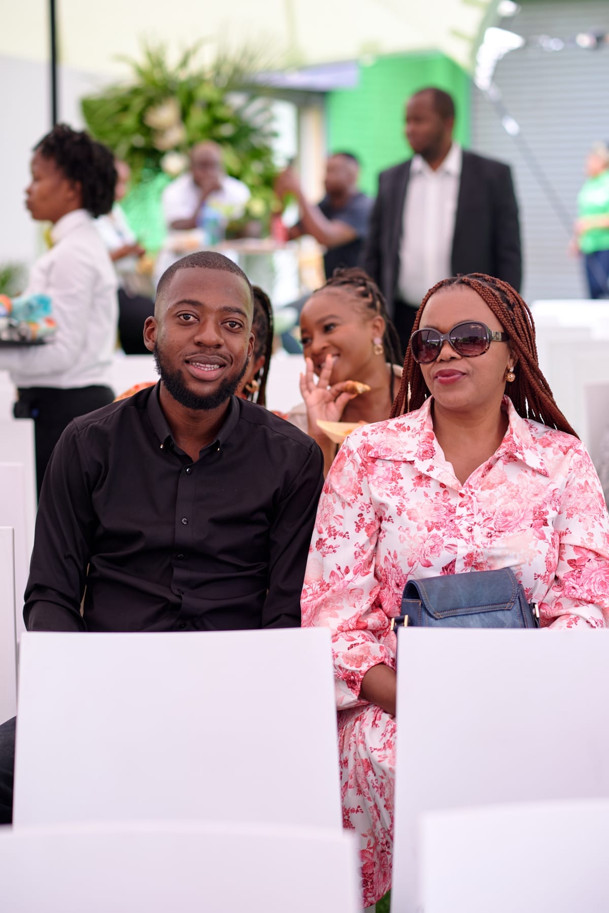 A man in a black shirt and a woman in a pink floral dress with plaits and sunglasses sit together smiling. They are seated indoors, with other people and greenery visible in the background. The mood is cheerful and relaxed.