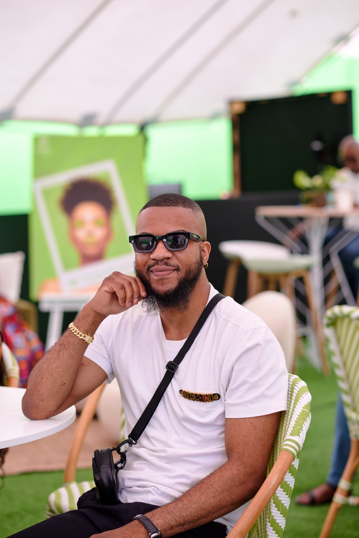 A man with a trimmed beard and sunglasses sits smiling at a round table. He wears a white T-shirt, gold bracelet, and crossbody bag. The background shows green decor, chairs, a bar table, and a blurred poster of a woman with an afro.
