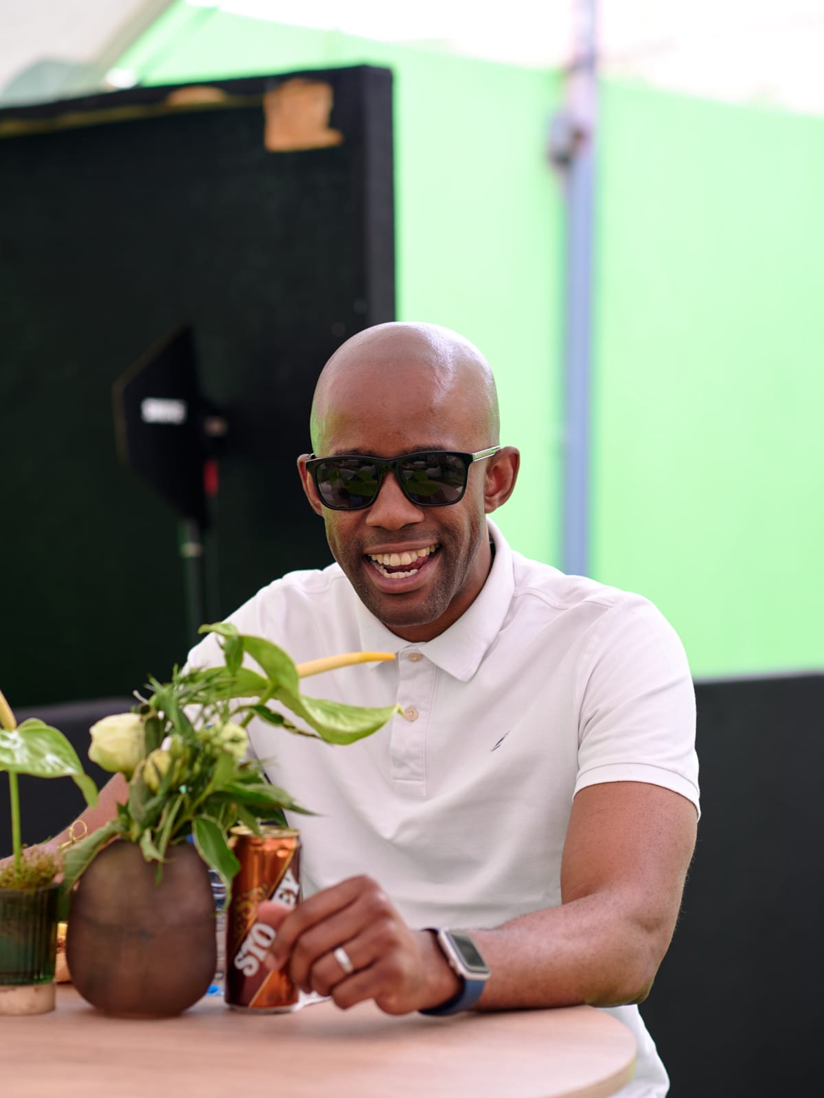 A smiling man wearing a white polo shirt and dark sunglasses sits at a round table. There are green and white flowers in a brown vase and a can of Stoney fizzy drink on the table. The background is green and black, with soft, natural light.
