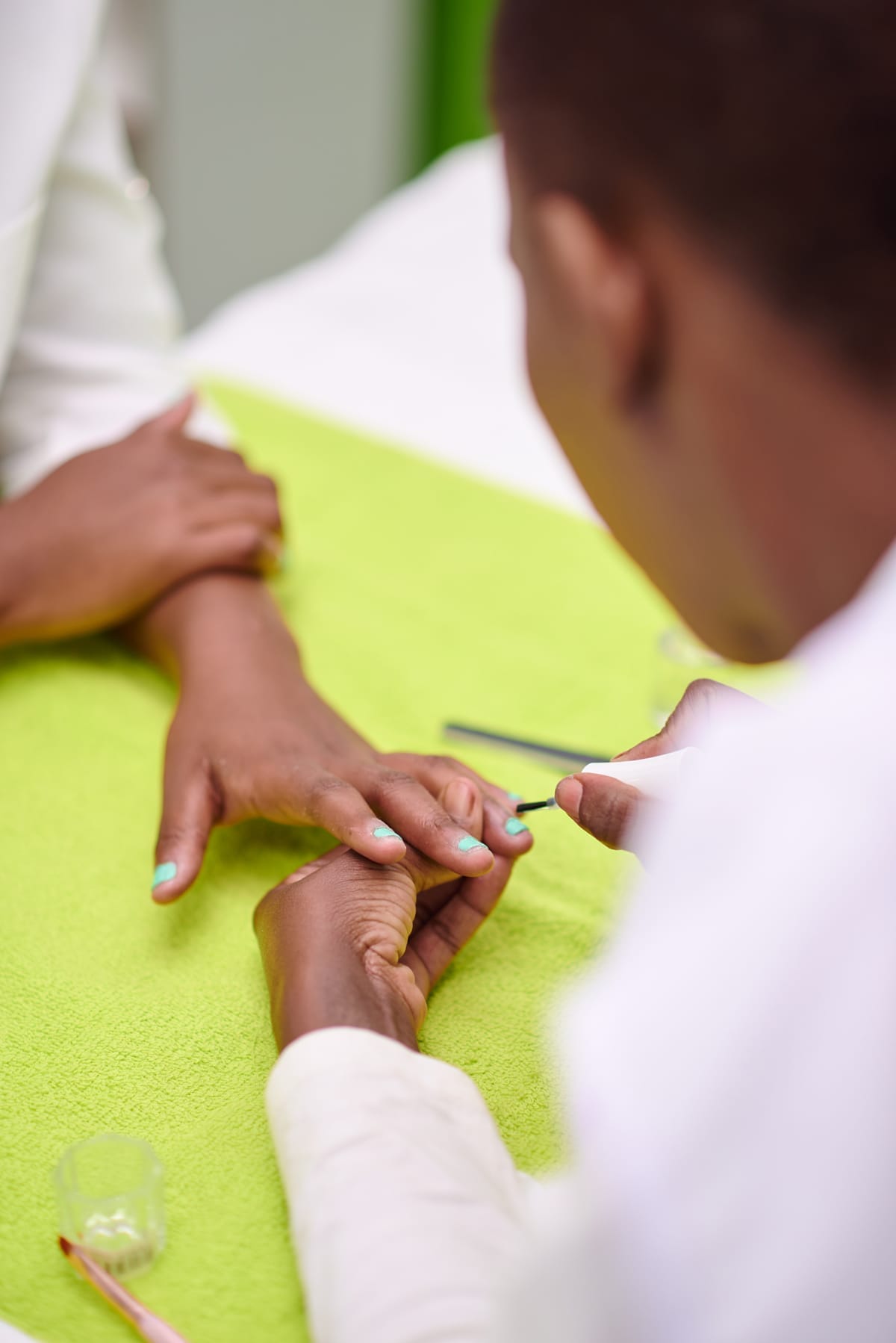 A person applies nail varnish to another person’s fingernails over a bright green towel, using a thin brush. The person receiving the manicure has light blue nail varnish on some fingers. Manicure tools and a small glass jar are nearby.