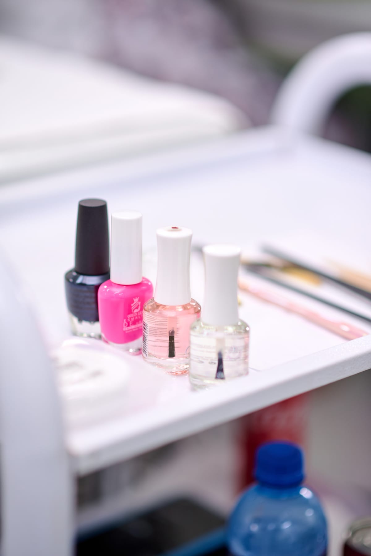 Four nail varnish bottles—one black, one pink, and two clear—are lined up on a white trolley. Behind them are nail files and brushes. A blue bottle top is partially visible at the bottom right corner. The scene is bright and clean.