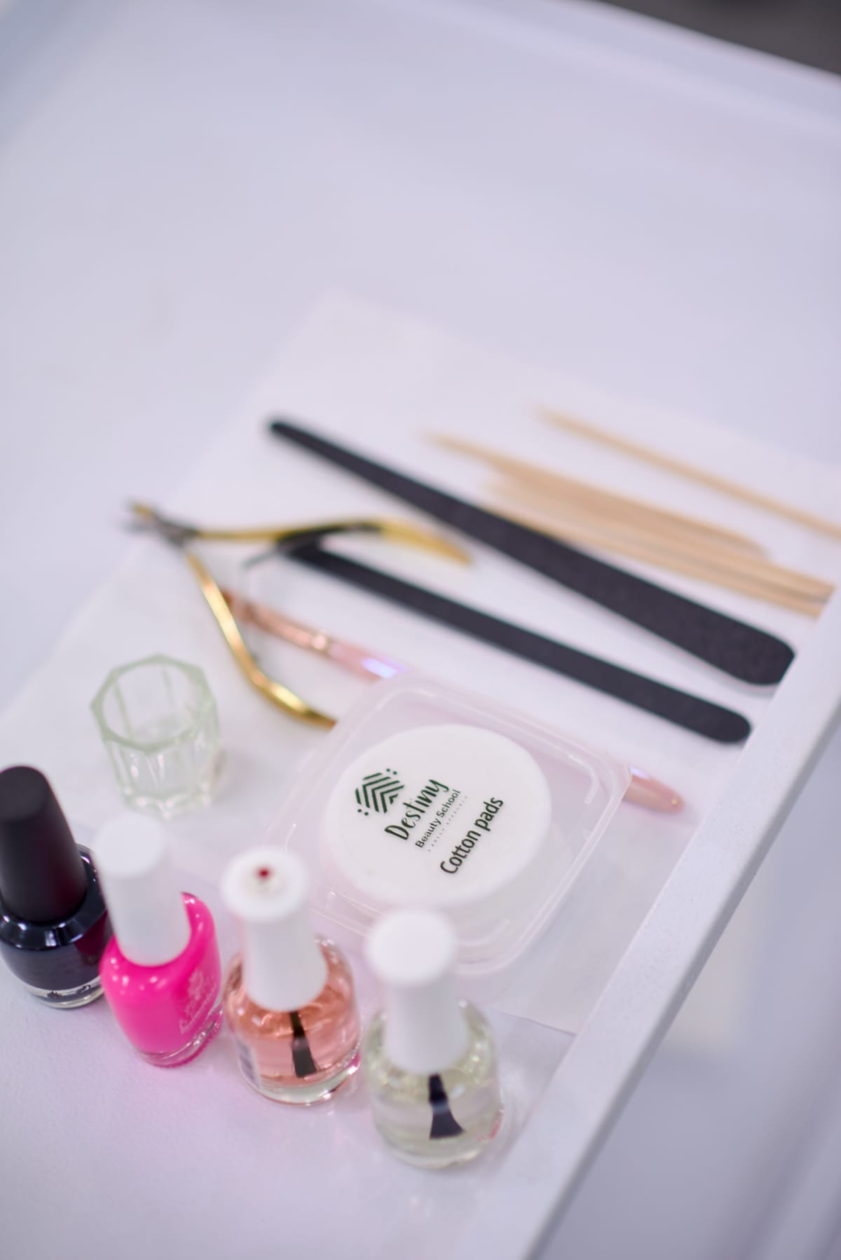 A close-up of a manicure set on a white tray, including nail polishes in pink, clear, and white, nail files, wooden cuticle sticks, cuticle nippers, a small glass holder, and a container labelled “Cotton Pads”.