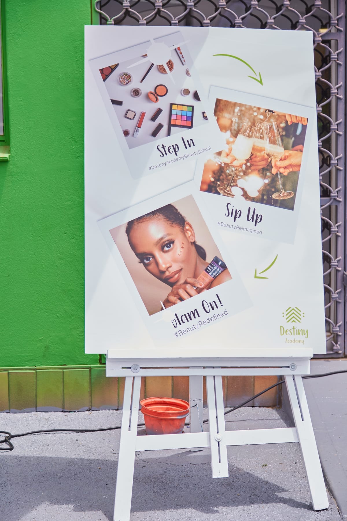 A large poster on an easel stands outside against a bright green wall. The poster features photos of make-up products, clinking glasses, and a woman with bold make-up. It includes the phrases “Step In,” “Sip Up,” and “Glam On!” with hashtags and the Destiny Academy logo.