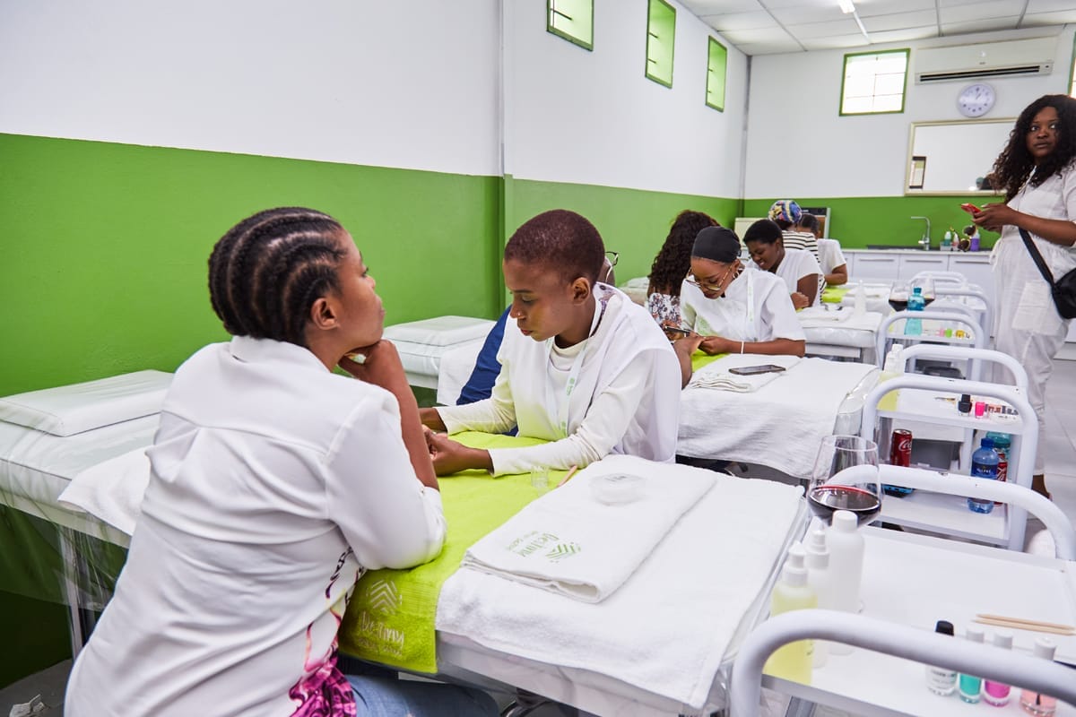 Several pairs of people, mostly women, sit at tables in a brightly lit classroom with green and white walls. They are engaged in manicure or nail care activities, wearing white uniforms. Nearby beds and supplies trolleys are visible, suggesting a beauty training setting.