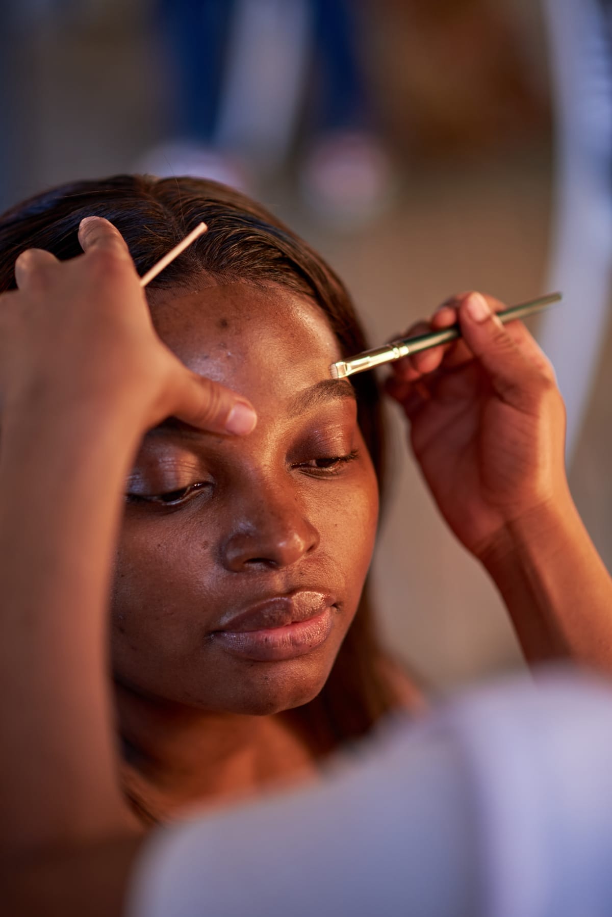 A close-up of a woman with closed eyes receiving eyebrow grooming. One hand holds a brush to her eyebrow whilst another hand uses a tool. The woman appears calm and relaxed, with soft lighting highlighting her face. The background is blurred.