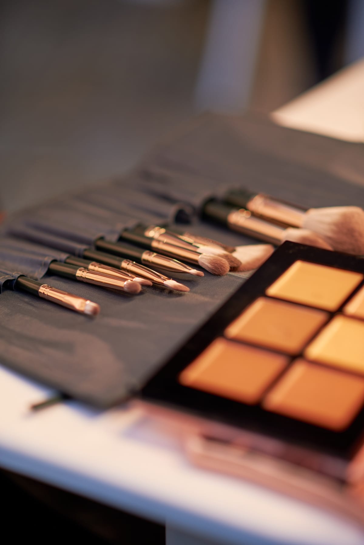 A close-up of make-up brushes with black and gold handles laid out on a grey cloth, alongside a make-up palette containing several shades of brown and gold powders, on a white surface. The background is softly blurred.
