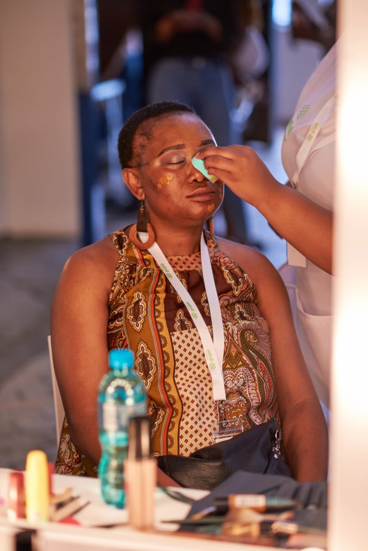 A woman sits with her eyes closed as another person applies make-up to her face using a green sponge. She wears a patterned sleeveless top and large hoop earrings, and has a white lanyard around her neck. Make-up items and a water bottle are on the table in front.