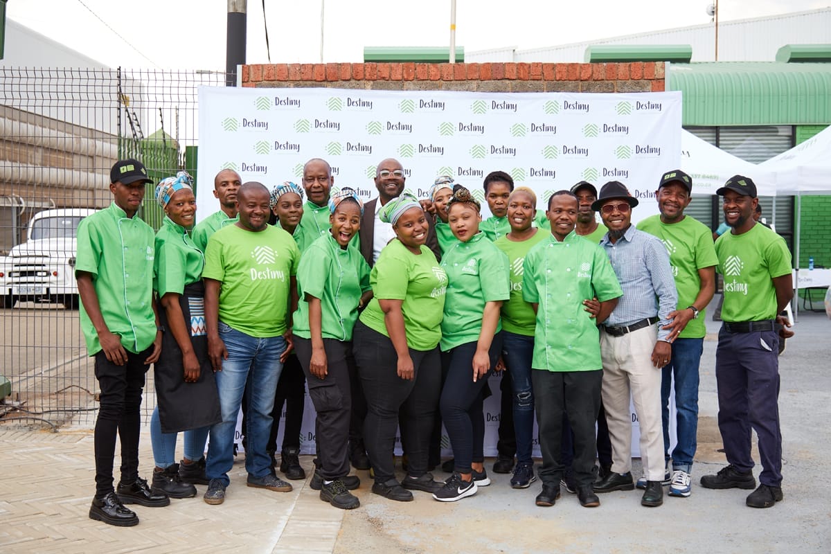A group of people stand together outdoors, smiling at the camera. They wear green shirts with 