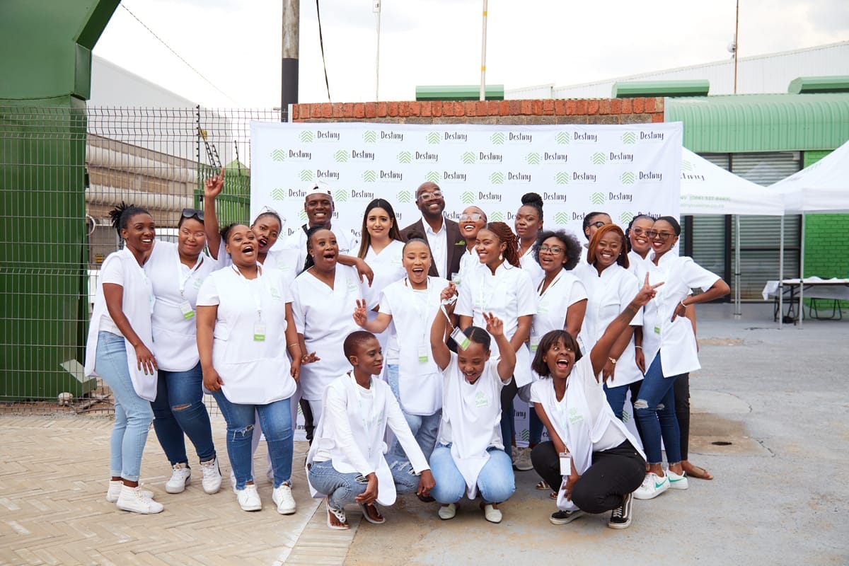 A group of smiling people in matching white shirts pose together outside in front of a branded backdrop. Many are making peace signs and enthusiastic gestures, conveying excitement and unity. The setting appears to be an outdoor event with modern buildings behind them.